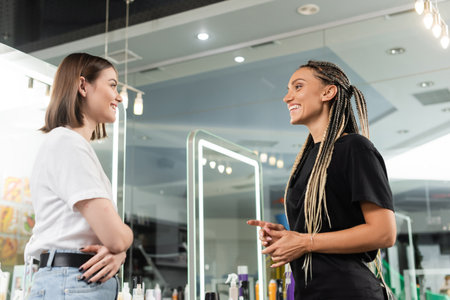 happy beauty worker welcoming female client, cheerful hair stylist with braids talking to smiling woman in salon, customer satisfaction, beauty industry, hair extension, hair colorist, salon jobの写真素材