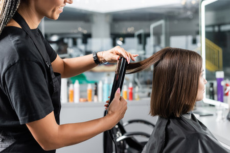 hair ironing, cheerful hairdresser with hair straightener styling hair of female customer, beauty worker, client satisfaction, brunette woman with short hair, beauty salon, hair fashionの写真素材