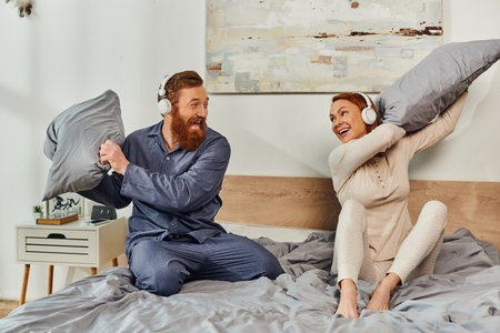 day off without kids, having fun, shared music, redhead husband and wife, happy couple in wireless headphones pillow fight, bearded man and carefree woman, tattooed music lovers, weekendsの写真素材