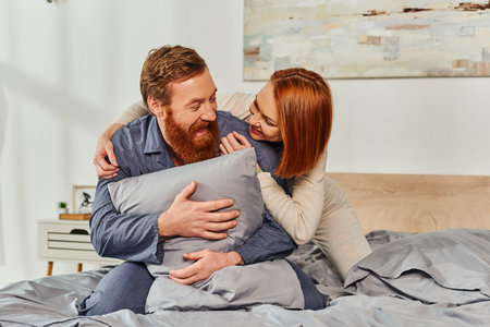 day off without kids, redhead woman hugging bearded husband with pillow, happy couple in sleepwear...