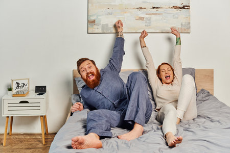 excited people, day off without kids, redhead and happy husband and wife, bearded man and woman with raised hands, parents alone at home, modern lifestyleの写真素材