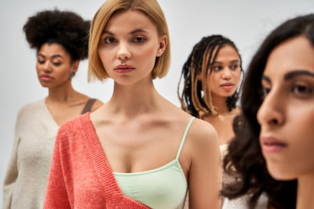 Portrait of young blonde woman in bra and wool jumper looking at camera while standing near blurred friends isolated on grey, different body types and self-acceptance, multicultural representationの写真素材