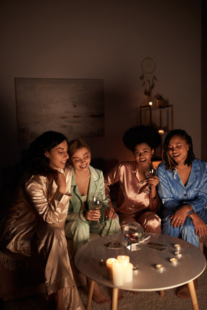 Positive multiethnic girlfriends in colorful pajama holding glasses of wine near tarot cards, crystal ball and candles during girls night at home, bonding time in comfortable sleepwear, divinationの写真素材