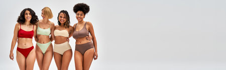 Laughing and confident multiethnic women in colorful lingerie hugging and posing together isolated on grey, different body types and self-acceptance, multicultural representation, bannerの写真素材