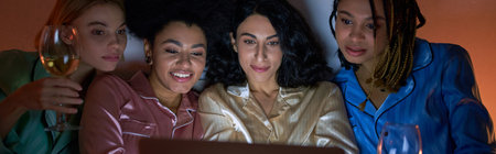 Cheerful and multiethnic women in colorful pajama using blurred laptop and holding glasses of wine during girls night at home, bonding time in comfortable sleepwear, bannerの写真素材
