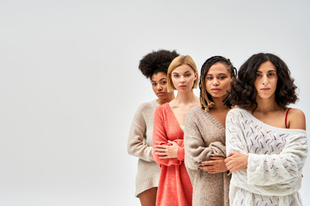 Multiethnic group of women in stylish knitted sweaters crossing arms and looking at camera isolated on grey, different body types and self-acceptance, multicultural representation, copy spaceの写真素材