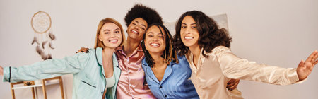 Positive multiethnic friends in colorful pajama hugging each other and looking at camera while having fun at home, bonding time in comfortable sleepwear, slumber party, bannerの写真素材