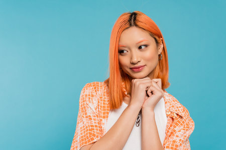 adorable, cheerful young asian woman with dyed red hair smiling and holding hands near face on vibrant blue background, pleased, generation z, casual attire, looking away, young cultureの写真素材