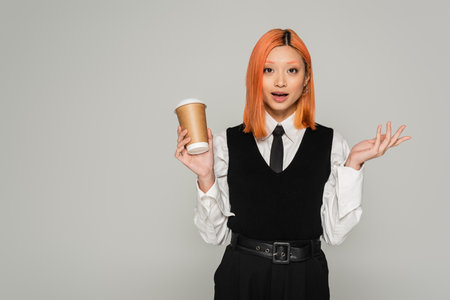 amazement, surprised asian woman with open mouth holding paper cup, gesturing and looking at camera on grey background, business casual style, white shirt, black tie and vest, colored red hairの写真素材