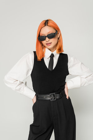 trendy asian fashion model in dark sunglasses, white shirt, black tie, vest and pants posing with...