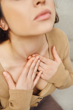 High angle view of young brunette woman in casual jumper touching neck during self-massage of thyroid gland in blurred house, self-care ritual and holistic wellness practices concept, tension reliefの写真素材