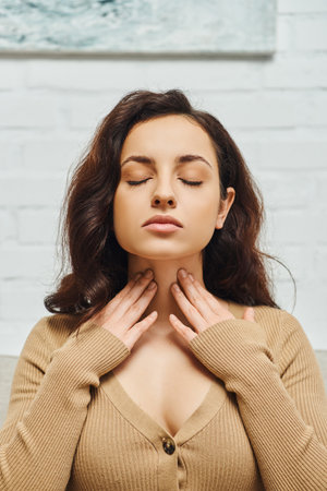 Portrait of young brunette woman in brown jumper checking thyroid gland on neck while sitting with closed eyes on couch in living room at home, focus on self-care and well-being conceptの写真素材