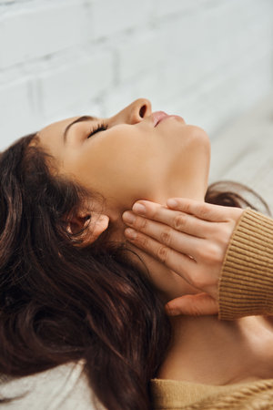 Young brunette woman in brown jumper touching lymphatic node on neck and relaxing during self-massage on couch at home, self-care ritual and holistic healing concept, tension reliefの写真素材