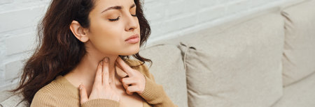 Young woman in casual jumper massaging lymphatic nodes on neck during wellness routine on couch at...