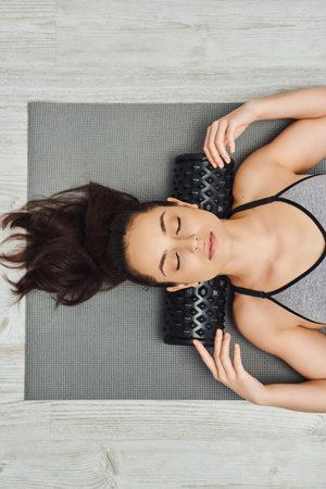 Top view of young relaxed brunette woman in sportswear using roller massager on neck while lying on fitness mat at home, home-based massage and holistic practices concept, myofascial releaseの写真素材