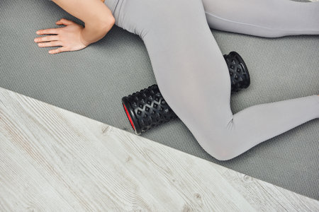 Cropped view of young woman in activewear massaging leg with roller massager and lying on fitness...
