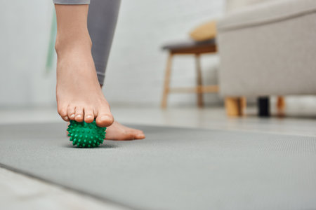 Cropped view of woman massaging foot with manual massage ball and standing on fitness mat at home, body relaxation and holistic wellness practices, balancing energyの写真素材