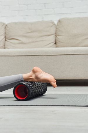 Cropped view of barefoot woman massaging leg with modern roller massager while lying on fitness mat near living room at home, promoting lymph flow and wellness at home concept, tension reliefの写真素材
