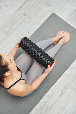 Overhead view of young barefoot woman in activewear holding roller massager and sitting on fitness mat in living room, home-based massage and holistic practices concept, myofascial releaseの写真素材