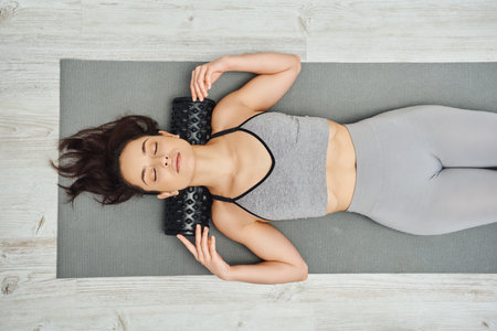 Top view of young brunette woman in sportswear using roller massager on neck while lying on fitness...
