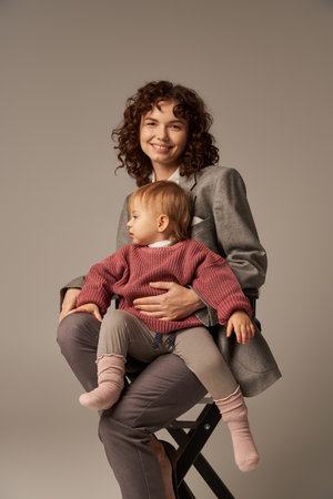 balanced lifestyle, career and family, curly woman in suit sitting on chair with child on grey background, professional success, motherhood, multitasking, quality time, work life balance conceptの写真素材