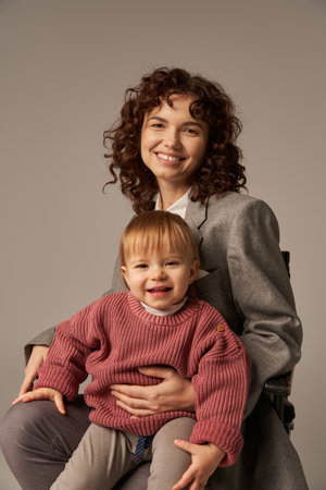 balanced lifestyle, growth and family, happy businesswoman sitting on chair with child on grey background, professional success, motherhood, multitasking, quality time, work life balance conceptの写真素材