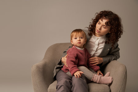 balancing work and life concept, curly woman in suit sitting in armchair with toddler daughter, grey background, multitasking, quality family time, mother and child, modern working parentの写真素材