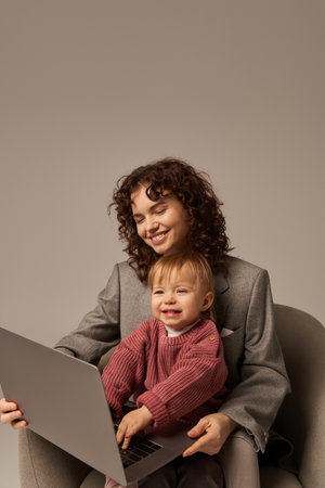 building successful career, balanced lifestyle, working parent, happy curly woman using laptop and sitting on armchair with daughter, mother and child, motherhood, multitaskingの写真素材