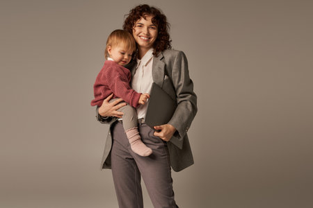 work life harmony, happy working mother holding laptop and toddler daughter on grey background, time management, self-confidence, modern parenting, professional success, businesswomanの写真素材
