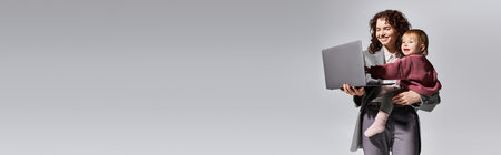 time management, happy working mother holding laptop and toddler daughter in arms on grey background, work life harmony, career and family, modern parenting, professional success, bannerの写真素材