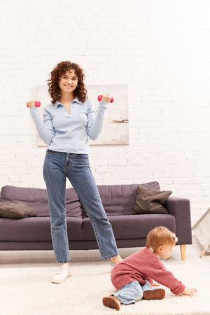 time management, working mother, balanced lifestyle, curly woman exercising with dumbbells near toddler daughter in cozy living room, home workout, sport, busy mom, physical activityの写真素材