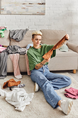 tattooed woman in casual style clothes holding suede boot while sorting belongings and thrift store...