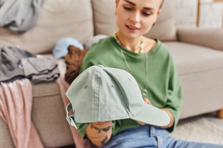 sorting clothes, young woman holding stylish cap during wardrobe items decluttering near couch at home, blurred background, sustainable living and mindful consumerism conceptの写真素材