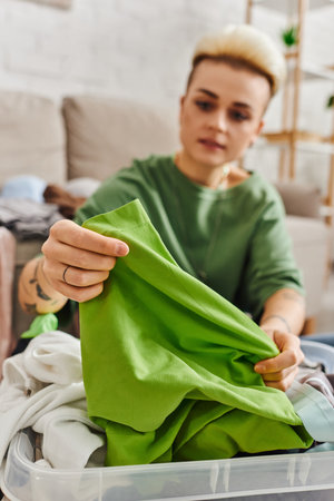 young tattooed woman reducing wardrobe, sorting clothes and holding green garment in living room at home, blurred background, sustainable living and mindful consumerism conceptの写真素材