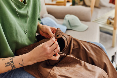 cropped view of young tattooed woman buttoning leather pants while sitting on couch near wardrobe items during decluttering process, sustainable living and mindful consumerism conceptの写真素材