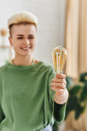 cheerful woman with trendy hairstyle, in casual clothes smiling and standing with energy saving light bulb at home on blurred background, sustainable lifestyle and environmentally conscious conceptの写真素材