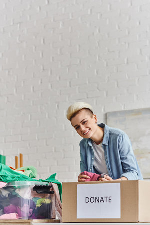young and joyful woman with trendy hairstyle holding pre-loved clothes and looking at camera near...