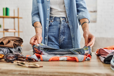partial view of young and tattooed woman in casual clothes sorting wardrobe items on table in living room, exchange market, eco-friendly swaps, sustainable living and mindful consumerism conceptの写真素材