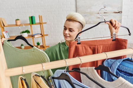 positive young woman holding hangers with pants and jumper near rack with wardrobe garments,...