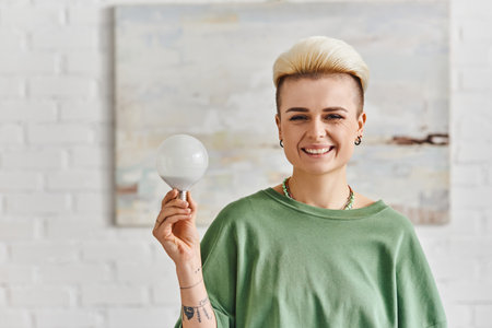 ecological efficiency, joyful tattooed woman with trendy hairstyle holding energy saving light bulb...