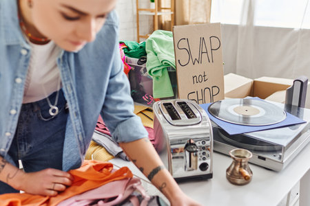 young tattooed woman sorting second-hand clothes near electric toaster, cezve, vinyl record player...