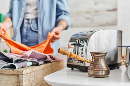 partial view of young woman sorting clothes near electric toaster and cezve, eco-friendly swaps, exchange market, blurred background, sustainable living and promoting circular economy conceptの写真素材