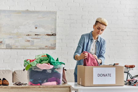donating for a cause, stylish tattooed woman packing garments in donation box near plastic container with second-hand items in living room, sustainable living and social responsibility conceptの写真素材