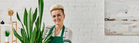 plant lover, young and cheerful woman with trendy hairstyle looking at camera near green and natural tropical plant in living room, sustainable home decor and green living concept, bannerの写真素材
