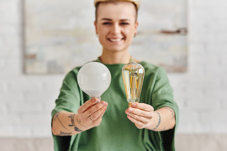 cheerful, young and tattooed woman in casual clothes showing energy saving light bulbs and looking at camera, environmentally conscious, sustainable lifestyle and environmentally conscious conceptの写真素材