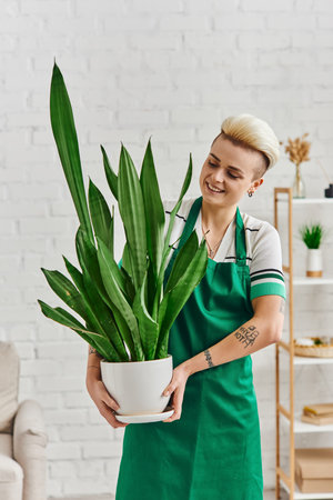 happiness, cheerful tattooed woman with trendy hairstyle holding flowerpot with exotic foliage plant in modern living room, eco-conscious lifestyle, sustainable home decor and green living conceptの写真素材