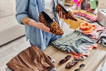 ethical consumption, exchange, cropped view of young tattooed woman holding stylish animal-print shoes near sunglasses and garments on table, sustainable living and mindful consumerism conceptの写真素材