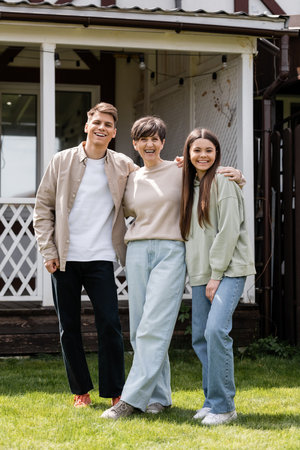 Full length of smiling middle aged mother hugging daughter and son while looking at camera during parents day celebration at backyard of house, parent-child relationship conceptの写真素材