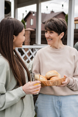modern parenting, celebration of parents day, happy middle aged mother holding buns and talking with her teenage daughter, modern parenting, suburban life, summer, family houseの写真素材
