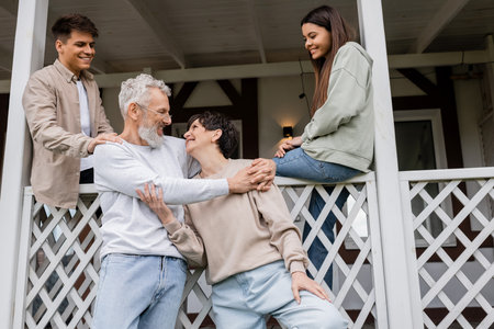 parents day, joyful siblings looking at middle aged parents hugging and smiling on porch of summer house, family celebration, bonding, moments to remember, modern parenting, happy marriageの写真素材